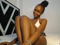 BlueBreeze, dark-skinned woman, sitting on bed, legs up, wearing pink bikini top, ass prominent, braided hair in bun, smiling, bracelet on right wrist, head against black-and-white headboard.