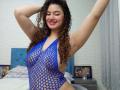 SanetPerlas with curly brown hair, wearing a blue fishnet top that reveals her tits and pussy, arms raised, smiling with pink lipstick in a bedroom.