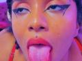 MeganWilliams with dark curly hair, red lipstick, black winged eyeliner, nose piercing, and tongue sticking out, wearing a red strap. Her face is slightly flushed with purple lighting.