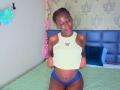 KharlaQueen with dark skin, small butterfly tank top, blue panties, braided hair, standing on bed, lifting top, showing flat stomach, small breasts, and pink lipstick.