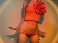 FlyFreedom with orange crop top, huge red bow, red panties, pulling them down, showing her flat stomach and bare pussy, blonde hair, shadow on wall, warm lighting.