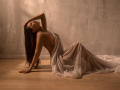 KarlaSoto with long brown hair, sideboob, and a sheer, flowing dress, sitting on a wooden floor, hand in hair, eyes closed, back arched, looking sexy.