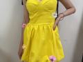 MiraMixi wearing a bright yellow dress with white and pink flower decals, showing her tits and hand with pink nail polish, standing against a plain wall.