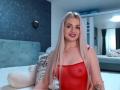 CoquineElise avec de longs cheveux blonds, un haut rouge transparent, des mamelons visibles, souriante, portant un collier avec une croix, assise sur un lit avec un animal en peluche blanc et une tête de lit bleue..