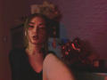 LelaTraverso with pouty lips, dark makeup, and blonde hair, wearing a black top, has her knee up, showing a necklace, in dim, red-tinted lighting.