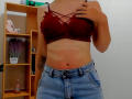 Steisii's red lace bra with criss-cross straps, showing her tits, and blue denim skirt, has a tan line and tattoo on her right arm, holding her bra strap.