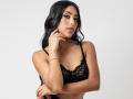 EmmaEstreilla with long black hair, wearing black lace bra, gold necklace, and bracelet, touching chin, looking at camera, arms crossed, showing her tits, pink lipstick, white background.