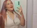 AntoniaBrunni with long red hair, red lipstick, white sleeveless shirt, holding green phone, taking mirror selfie, showing medium-sized boobs, smiling, in bathroom, holding phone with white nails.
