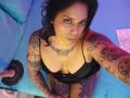 Chili with tattoos, black lingerie, and cleavage, looking up, sitting on blue couch, pink dildo on black ring, laptop on right, dark hair, blue eyes, purple and blue lighting.