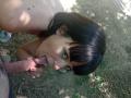 OrianaAndFriends, with dark hair and bangs, is fellating a man's erect, uncircumcised penis. Her pink-nail-holding hand grips his cock, eyes looking up. She's topless, on grass.