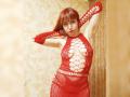 LaylaHottyX with red hair, wearing a red, see-through, lace-up dress, exposing her tits and pussy, has arms raised, standing in a room with beige, patterned wallpaper.