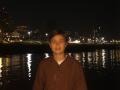 MarybelleDoerr, with short brown hair, smiling at night, wearing a brown button-up shirt, standing by a reflective water surface, city lights in the background.