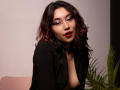 AliCipher with wavy dark brown hair, red lipstick, black shirt open showing cleavage, gold necklace, and subtle makeup, looking slightly to the side with a confident expression.