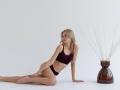 EolantaMarina with small tits and a slim ass, wearing a dark purple bra and black panties, sits on a white floor with a brown vase of white branches.
