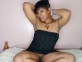 RareGem with dark skin, short black hair, arms raised, wearing a black strapless dress, exposing her pussy and armpits, sitting on a bed, wearing a blue bracelet.