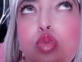 AlessadraJoness's face with pink blushed cheeks, pouty red lips, silver hair, nose ring, and looking up with blue eyes, wearing pink earrings. Her face is centered, close-up.