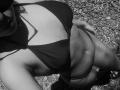 black-and-white photo of AlexXas, showing her tits in a dark bikini top, pebbled ground below, shadow on chest, bracelet on right wrist, partial face with hat.
