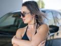 Milagres with dark sunglasses, black halter top, and hoop earrings, arms crossed, showing her tits, long gray-streaked hair, and a black choker. Car in the background.