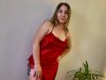 Aminas with medium tits, wearing a red satin dress, standing, hand on hip, slight lean, blonde hair, tattoo on thigh, green plant in bottom right, smiling.
