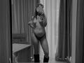 Black-and-white mirror selfie of AlexXas with glasses, topless, showing tits, wearing dark panties and boots, holding phone, standing in room with curtains and wooden furniture.