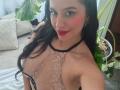 MadissonFontaine with long black hair, red lipstick, and pink cheek blush, wearing a sheer lace top, exposing her big tits, smiling at the camera, indoors, with a potted plant.