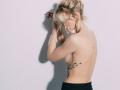 Topless EolantaMarina with blonde hair in a messy bun, black pants, side tattoo, hands in her hair, shadow on white wall. Her tits are small and she's looking down.