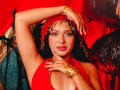 LilyConorr with red headscarf, gold jewelry, and red halter top, showing off her tits and shiny lips, arm raised, green eyes, and black hair, against red background.