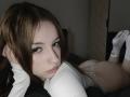TaniaRaffa with brown hair, lying on bed, white long-sleeve top, lace white panties, showing her ass, white socks with red logo, looking at camera, dark room.
