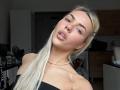 ArnettaNazario has long blonde hair, pouty pink lips, and a pearl necklace. She's wearing a black off-shoulder top, with her tits slightly visible. Her ass is not shown.