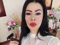 SofiMontes has black hair, thick eyebrows, pink eyeshadow, red lipstick, and is wearing a sheer white top with lace patterns. Her pink lips are slightly puckered.