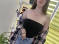 Nesie with bare boobs in a black strapless top, pink plaid shirt off her shoulders, and blue jeans, standing in front of a wall with butterfly stickers and a toy.