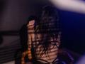IngaDrein's back is to the camera, sitting in a wooden chair, wearing black strappy lingerie, with long dark hair and shadowed stripes from blinds, in a dimly lit room.