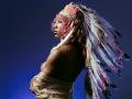 DaniaMarcell with pink curly hair, wearing a Native American-style feather headdress, tan off-shoulder lace top, holding her tits, tattoos on right arm, against a dark blue background.