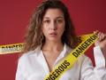 Sanella with curly brown hair, wearing a white shirt, holding "Danger" and "Genuine" tape, has a serious expression, and her tits are slightly visible under the shirt.