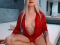 CoquineElise, avec ses longs cheveux blancs, son rouge à lèvres rouge et sa robe rouge transparente qui révèle ses seins, et un petit pendentif en forme de croix, assise sur un lit, les jambes croisées, avec un tatouage sur le bras droit..