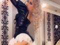 MademoiselleBella with blonde curls, black leather top, and white ruffled skirt, showing her bare ass, standing in a room with black and white damask wallpaper.