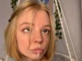 MyrleMogush has blonde hair, blue eyes, and a small amount of white fluid dripping from her slightly parted, shiny pink lips. She has a few freckles on her face.
