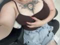 DiLaria with big tits, brown tank top, red nails, sun face tattoo, and frayed denim shorts, holding her chest on a gray chair.