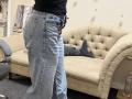 Dalaila's ass in blue, slightly flared, high-waisted jeans standing in front of a beige tufted couch with a gray pillow. Her black top is visible. Her feet are off-camera.