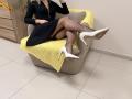 Rorria, vêtue d'une robe noire, avec des bas résille et des talons blancs, les jambes croisées, assise sur une chaise recouverte de jaune, son fessier légèrement exposé, dans une pièce avec des meubles en bois..