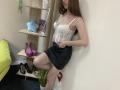 StellaNoir with long brown hair, white lace corset, showing her small tits, black skirt, leg raised, pink heels, black heels, wooden shelf with shoes, green couch, potted plant.