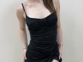 Ramilla wearing a black, ruched, sleeveless dress, showing her small tits, with a gold wristwatch on her right hand, her left hand on her dress's side.