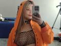 Melani Becker, met lang blond haar, draagt een oranje hooded jas en een zwarte nethemd, waarbij haar borsten zichtbaar zijn. Ze houdt een telefoon vast en maakt een selfie in de spiegel, in een slaapkamer..