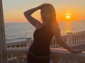 LeScharwath stands in a black dress, hand in hair, against a sunset ocean view, with a white balcony and gold chain belt, looking sexy and confident.