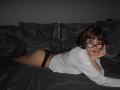 ErynAttridge lies on a bed, face resting on her hand, wearing black panties and a white shirt. She has short brown hair, glasses, and a seductive, half-smiling expression. Dark pillows behind.