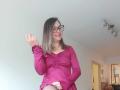 ArianaBlonde with glasses, smiling, wearing a pink dress, raising her right hand, showing her tits slightly, in a room with a ceiling light.
