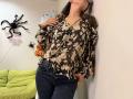 Tilliana, com os seios à mostra sob uma blusa floral marrom, calças jeans escuras e óculos, apoiada em uma parede, com a mão no peito, em um quarto com decoração de Halloween..