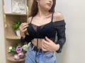 Rioria with big tits, wearing a black sheer off-shoulder top and blue jeans, holding her top, showing a black bra, gold earrings, and a pink lip.