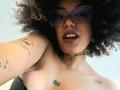 ScarlethVictoria with curly brown hair, wearing glasses, choker, and dark lipstick, showing tattoos on arm and chest, with a slight open-mouth smile, and small visible breasts.