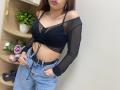 Rioria with her tits in a sheer black top, blue jeans, and hands in pockets, standing next to a wooden shelf with flowers and a potted plant.